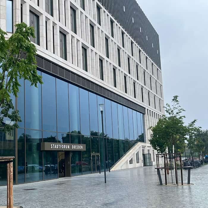 Außenansicht des modernen Gebäudes Stadtforum Dresden an der Waisenhausstraße mit großer Glasfront, hellem Stein, gepflastertem Vorplatz und jungen Bäumen unter bedecktem Himmel. 