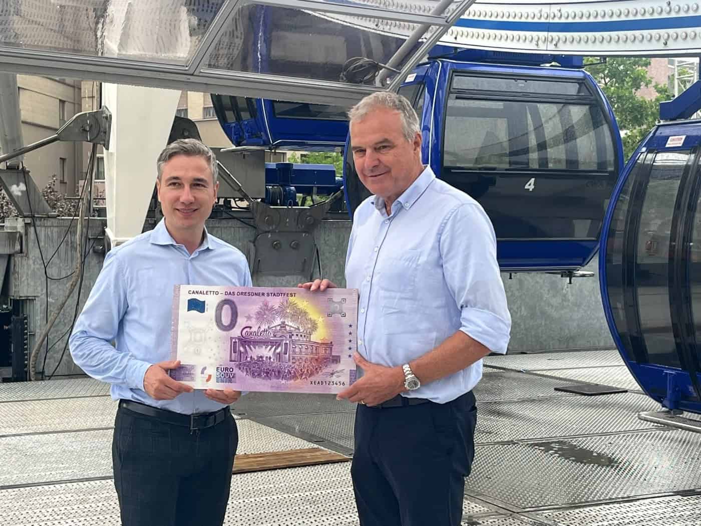 Baubürgermeister Stephan Kühn und Riesenrad-Betreiber Oscar Bruch halten gemeinsam einen übergroßen Null-Euro-Souvenirschein des Canaletto-Stadtfests vor blauen Riesenrad-Gondeln.
