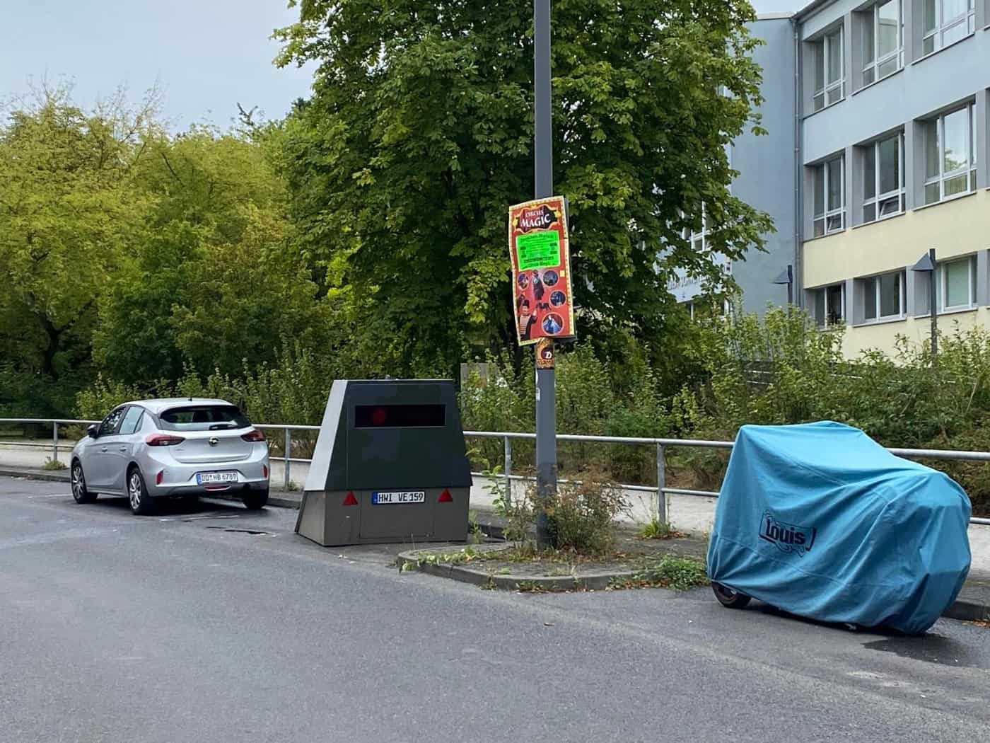 Blitzeranhänger HWI-VE 159 mit neuer dunkler Frontfolierung steht am Straßenrand der Georg-Palitzsch-Straße in Dresden-Prohlis direkt vor dem Gebäude einer Schule. Im Hintergrund stehen Bäume unter bedecktem Himmel, links parkt ein Auto, rechts ein verhülltes Motorrad.