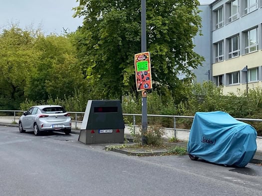 Blitzeranhänger HWI-VE 159 mit neuer dunkler Frontfolierung steht am Straßenrand der Georg-Palitzsch-Straße in Dresden-Prohlis direkt vor dem Gebäude einer Schule. Im Hintergrund stehen Bäume unter bedecktem Himmel, links parkt ein Auto, rechts ein verhülltes Motorrad.