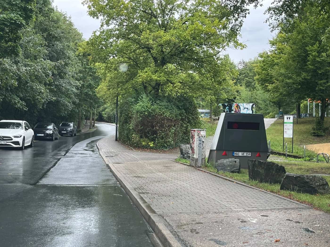 Ein getarnter Blitzeranhänger mit dem Kennzeichen HWI VE 159 steht auf einem nassen Gehweg an der Gamigstraße in Dresden. Rechts befinden sich Grünflächen und Felsen, links führt die feuchte Straße an parkenden Autos und Bäumen vorbei.