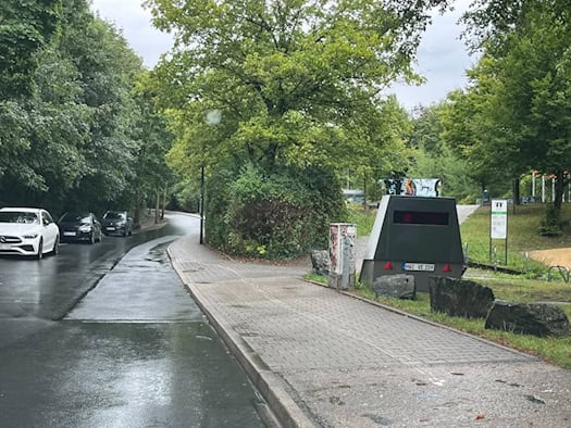 Ein getarnter Blitzeranhänger mit dem Kennzeichen HWI VE 159 steht auf einem nassen Gehweg an der Gamigstraße in Dresden. Rechts befinden sich Grünflächen und Felsen, links führt die feuchte Straße an parkenden Autos und Bäumen vorbei.
