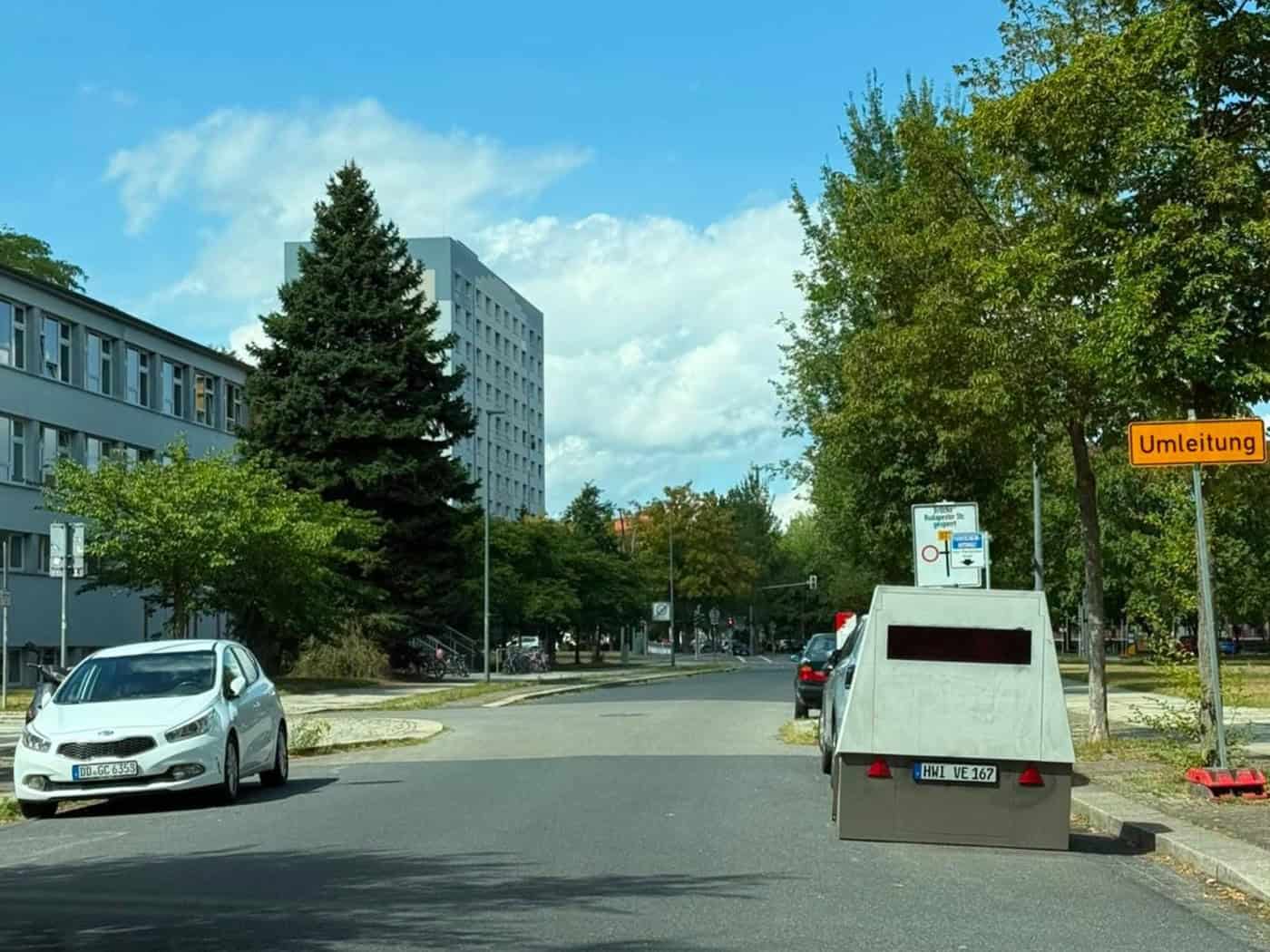Ein grauer Blitzeranhänger mit dem Kennzeichen HWI VE 167 steht am rechten Straßenrand der Josephinenstraße in Dresden. Rechts daneben steht ein gelbes Umleitungsschild, links ist ein Schulgebäude und ein weißes Auto zu sehen.