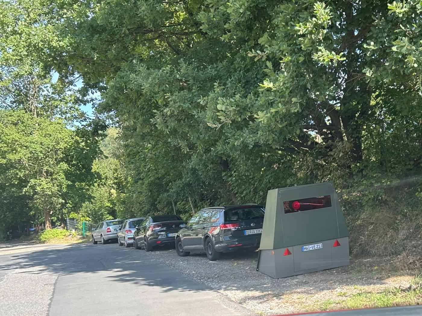 Blick aus einem Auto auf einen grauen gepanzerten Blitzeranhänger, der am schattigen Straßenrand der Tännichtstraße in Dresden unter Bäumen neben einem Metallzaun aufgestellt ist.