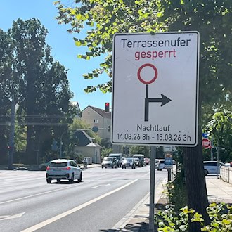 Ein weißes Verkehrsschild informiert über die Sperrung des Terrassenufers in Dresden. Der Aufdruck zeigt ein Umleitungssymbol sowie den Zeitraum „Nachtlauf 14.08.26 8h - 15.08.26 3h“. Im Hintergrund fahren Autos auf der mehrspurigen Straße.