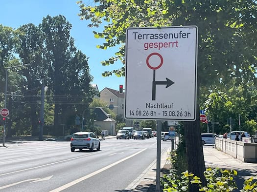 Ein weißes Verkehrsschild informiert über die Sperrung des Terrassenufers in Dresden. Der Aufdruck zeigt ein Umleitungssymbol sowie den Zeitraum „Nachtlauf 14.08.26 8h - 15.08.26 3h“. Im Hintergrund fahren Autos auf der mehrspurigen Straße.