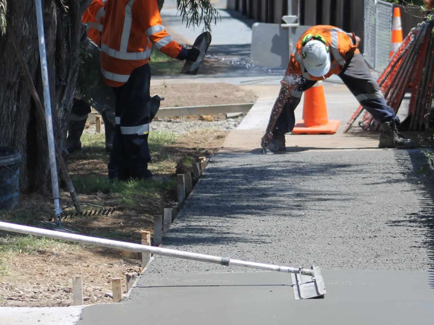 Bauarbeiter in orangefarbener Warnkleidung sanieren einen Gehweg. Einer glättet die frisch betonierte Fläche mit einem Werkzeug, im Hintergrund steht ein Betonmischer.