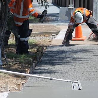 Bauarbeiter in orangefarbener Warnkleidung sanieren einen Gehweg. Einer glättet die frisch betonierte Fläche mit einem Werkzeug, im Hintergrund steht ein Betonmischer.