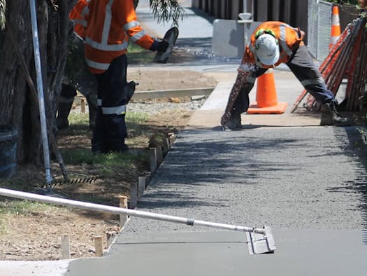 Bauarbeiter in orangefarbener Warnkleidung sanieren einen Gehweg. Einer glättet die frisch betonierte Fläche mit einem Werkzeug, im Hintergrund steht ein Betonmischer.