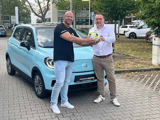 Stefan Lasch (links) und Christian Schleicher halten gemeinsam einen Fußball vor einem hellblauen Elektroauto auf dem Firmengelände des Autohauses Dresden. Beide lächeln in die Kamera.