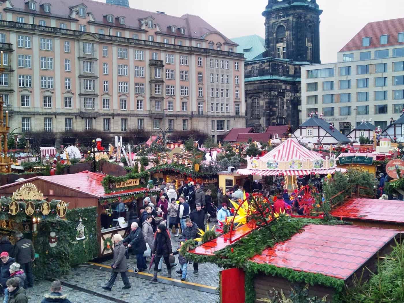 Blick von oben auf den belebten Dresdner Striezelmarkt mit weihnachtlich geschmückten Holzbuden, einem historischen Karussell mit rot-weißem Dach und Besuchern vor der Kulisse historischer Gebäude in der Dresdner Innenstadt.