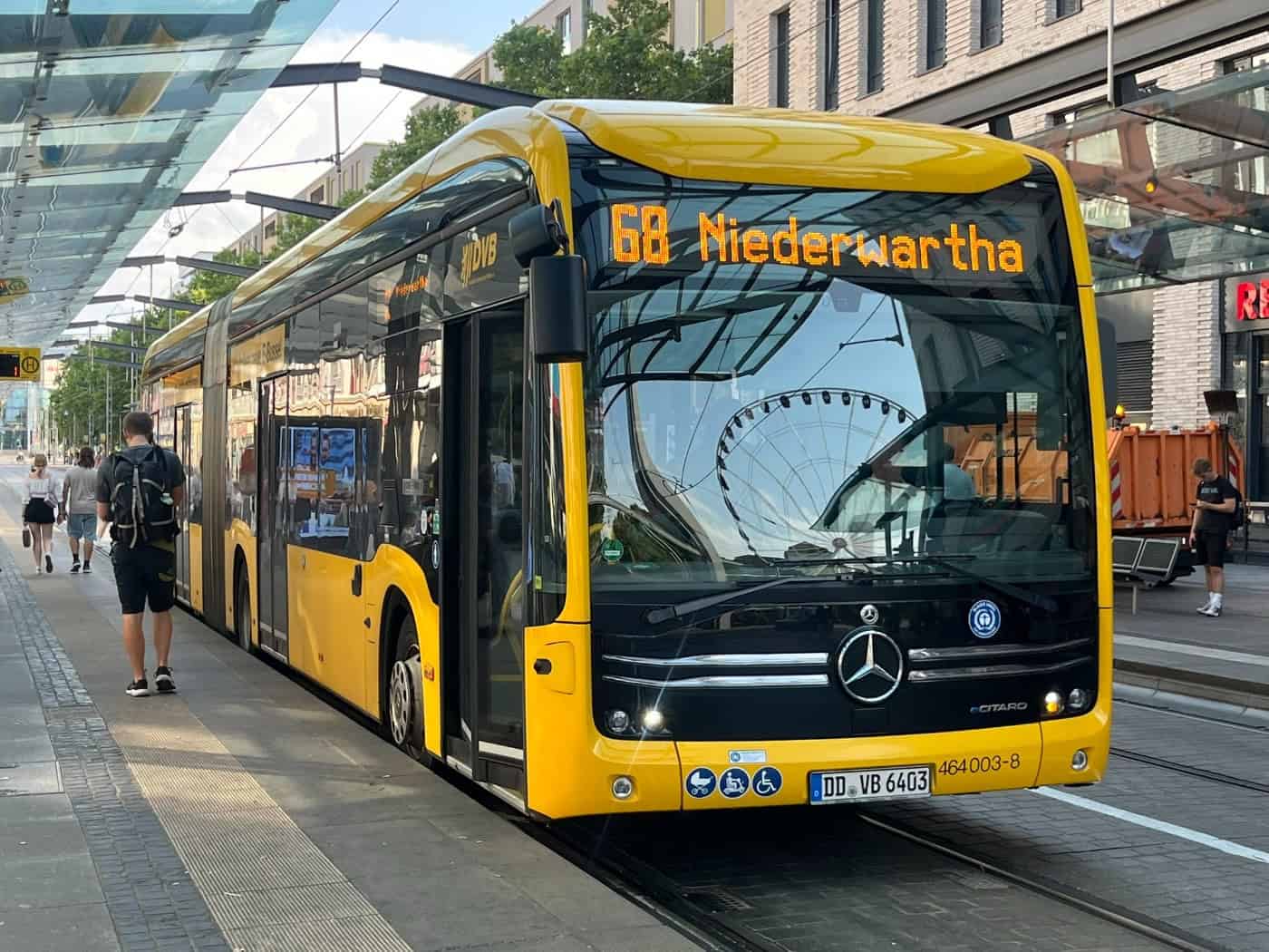 Ein gelb-schwarzer DVB-Elektrobus der Linie 68 mit Zielanzeige Niederwartha steht an einer überdachten Haltestelle. Auf der Windschutzscheibe spiegelt sich ein Riesenrad, links gehen Passagiere am Bahnsteig entlang.