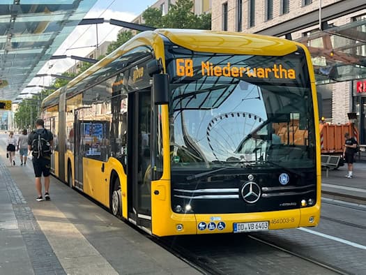 Ein gelb-schwarzer DVB-Elektrobus der Linie 68 mit Zielanzeige Niederwartha steht an einer überdachten Haltestelle. Auf der Windschutzscheibe spiegelt sich ein Riesenrad, links gehen Passagiere am Bahnsteig entlang.
