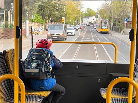 Blick aus dem Innenraum einer Dresdner Straßenbahn durch die Scheibe auf die Straße. Im Vordergrund sitzt ein Kind mit Helm und Schulranzen auf einem gelben Sitz, im Hintergrund kommt eine gelbe Straßenbahn entgegen.