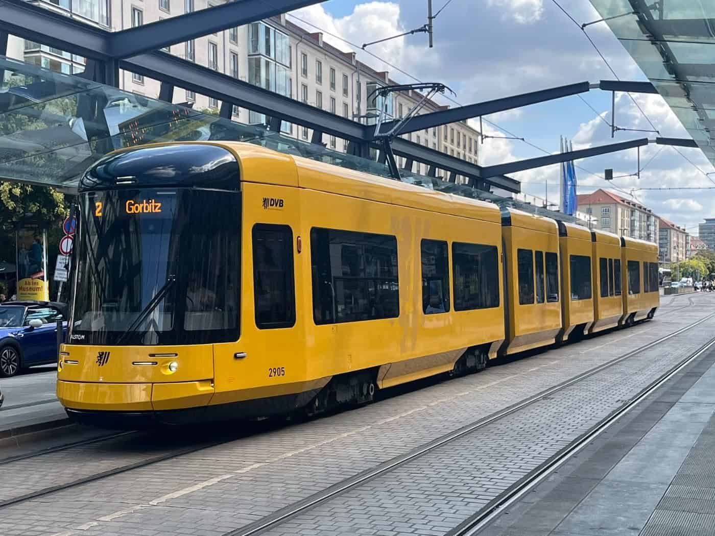 Eine moderne, gelbe DVB-Straßenbahn der Linie 2 mit dem Fahrtziel „Gorbitz“ steht unter einem Glasdach an einer Haltestelle in Dresden. Im Hintergrund gehen Passanten am Bahnsteig entlang bei weitgehend sonnigem Wetter.