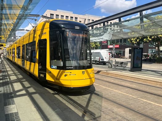 Eine gelb-schwarze Straßenbahn der Dresdner Verkehrsbetriebe (DVB) hält unter dem Glasdach der Haltestelle Postplatz. Die Digitalanzeige zeigt Linie 2 Kleinzschachwitz. Links stehen Fahrgäste auf dem Bahnsteig.
