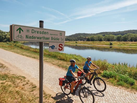 Zwei Frauen fahren auf E-Bikes mit Packtaschen auf dem gepflasterten Elberadweg am Flussufer entlang. Im Vordergrund steht ein grünes Schild mit Wegweisern nach Dresden und zur Niederwarthaer Brücke unter sonnigem blauem Himmel.