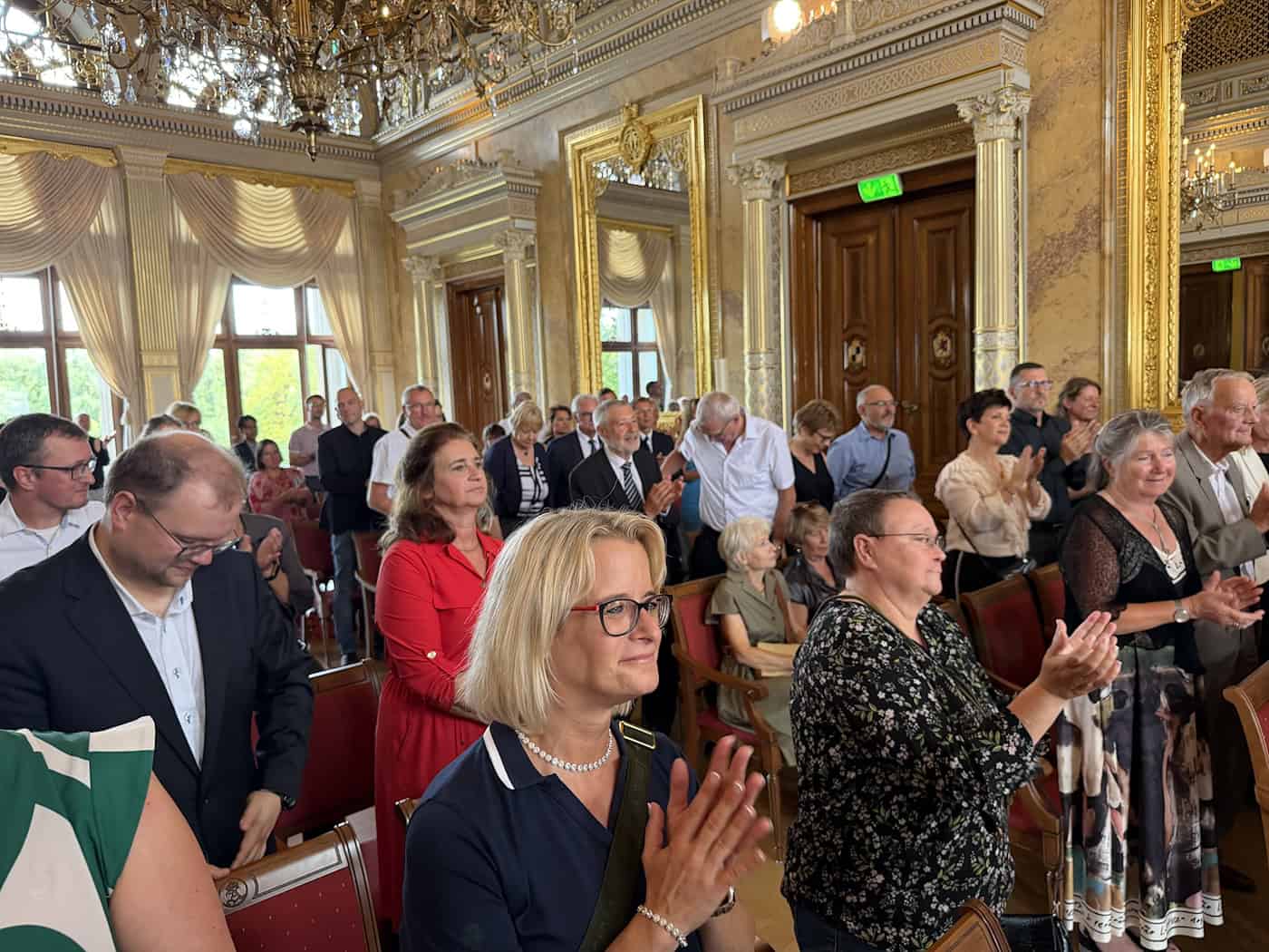 Blick in den gefüllten, golden dekorierten Festsaal von Schloss Albrechtsberg. Die Gäste im Publikum stehen und applaudieren lächelnd während der Verleihung des Erich-Kästner-Preises.