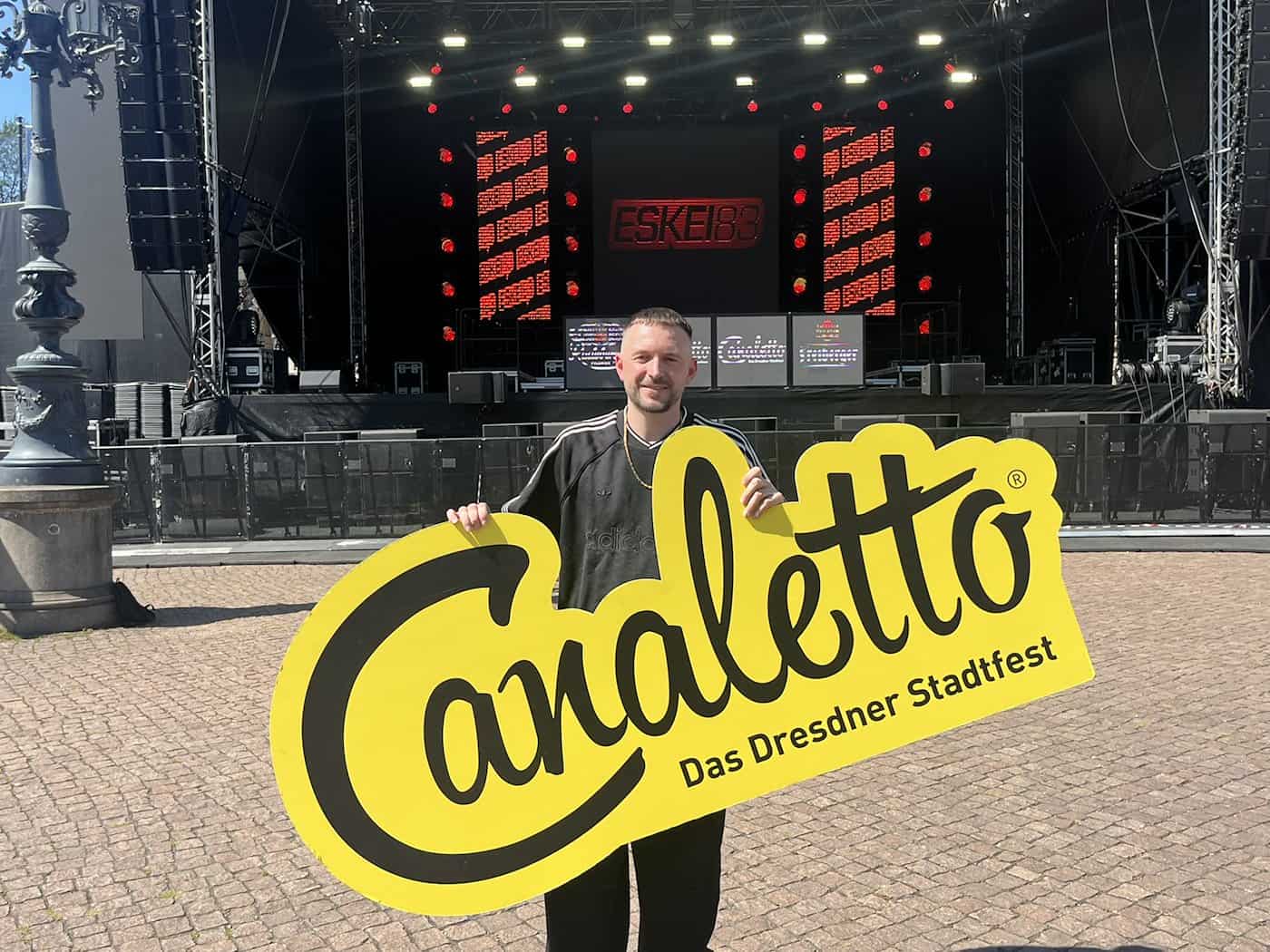 DJ Eskei83 steht vor der großen Stadtfest-Bühne auf dem Dresdner Theaterplatz und hält lächelnd ein großes gelbes Schild mit der Aufschrift „Canaletto - Das Dresdner Stadtfest“ in den Händen.
