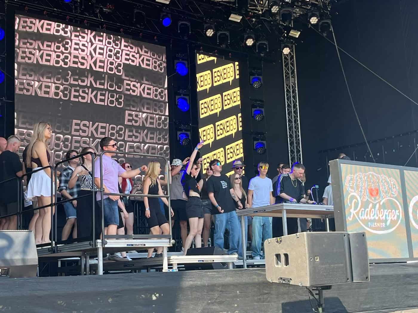 DJ Eskei83 legt auf einer großen Festivalbühne am DJ-Pult auf. Hinter ihm zeigen große Bildschirme seinen Namen, während eine Gruppe junger Leute auf der Bühne tanzt und feiert.