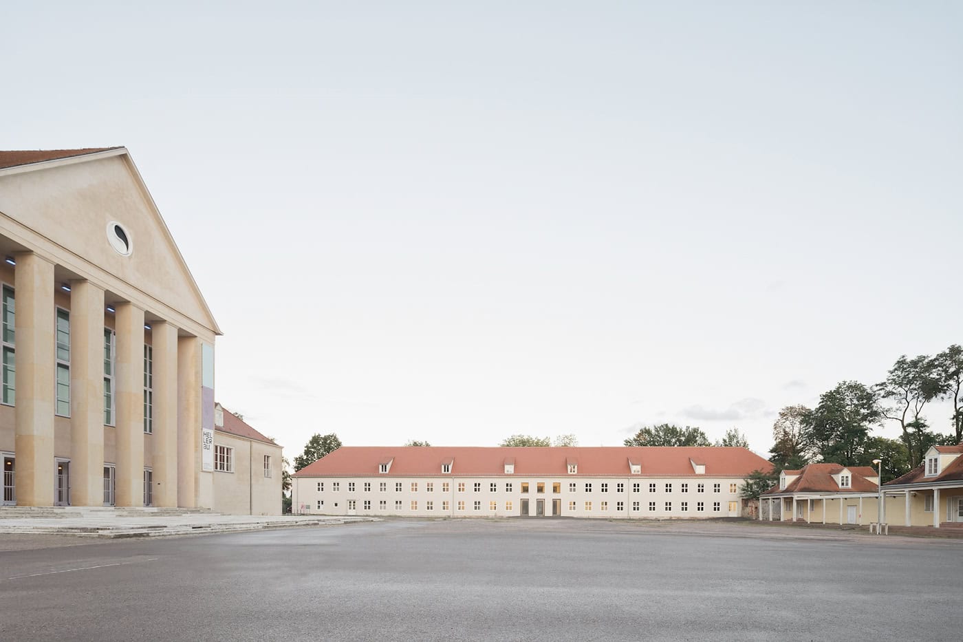 Eine Weitwinkel-Außenaufnahme des Festspielhaus Hellerau-Komplexes in Dresden. Im Vordergrund ein großer, leerer Platz. Links das Hauptgebäude mit Säulen und Portikus, in der Mitte der langgestreckte, sanierte Ostflügel mit rotem Ziegeldach. Heller Himmel.