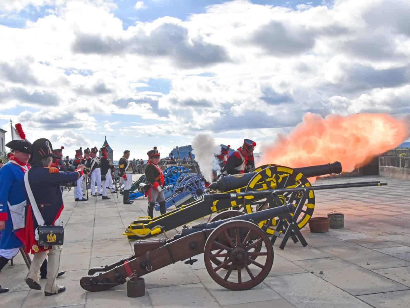 Historische Darsteller in Uniformen feuern eine gelb-schwarze Kanone auf einer Steinplattform der Festung Königstein ab. Dabei entsteht eine große orangefarbene Feuerwolke und Rauch unter leicht bewölktem Himmel.