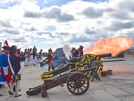 Historische Darsteller in Uniformen feuern eine gelb-schwarze Kanone auf einer Steinplattform der Festung Königstein ab. Dabei entsteht eine große orangefarbene Feuerwolke und Rauch unter leicht bewölktem Himmel.