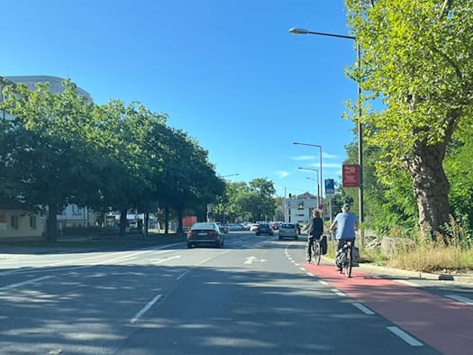 Blick auf die sonnenbeschienene Fetscherstraße in Dresden. Rechts fahren zwei Radfahrer auf einer rot markierten Radspur neben mehreren Autofahrspuren.