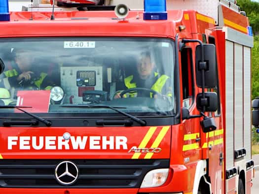 Ein rotes Feuerwehrfahrzeug fährt mit Blaulicht auf einer Straße. In der Fahrerkabine sind Einsatzkräfte in Schutzkleidung zu sehen.