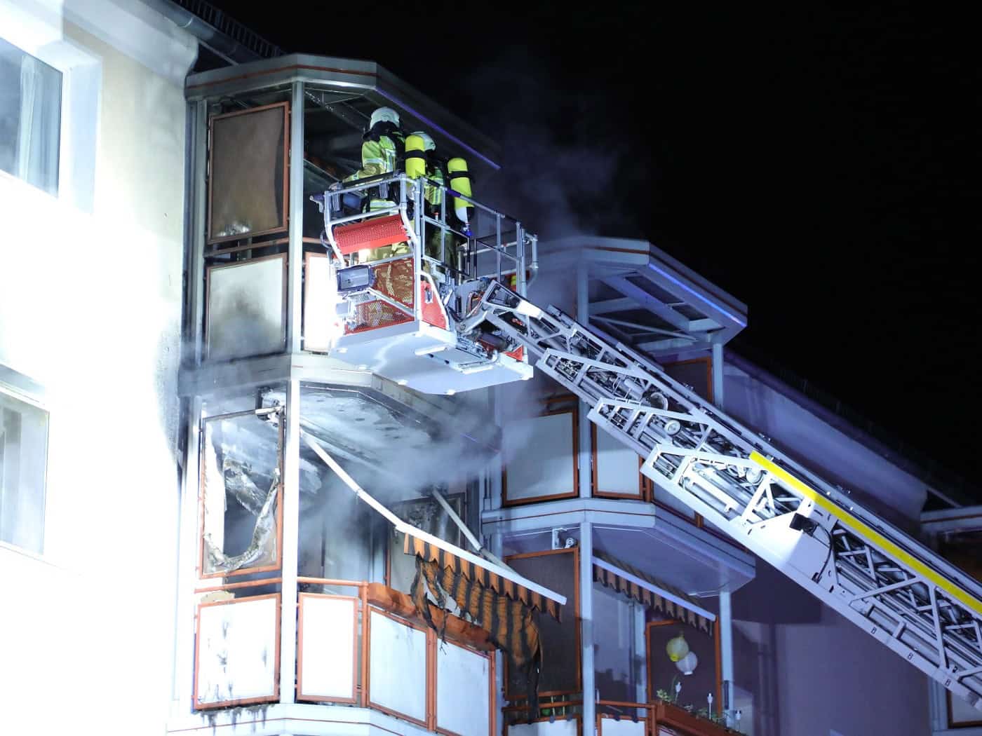 Feuerwehrleute in Schutzkleidung auf einer Drehleiter löschen einen Brand an einem Wohnungsbalkon im zweiten Obergeschoss eines Mehrfamilienhauses bei Nacht. Rauch steigt auf, Teile der Balkonverkleidung und Markise sind sichtbar verkohlt.