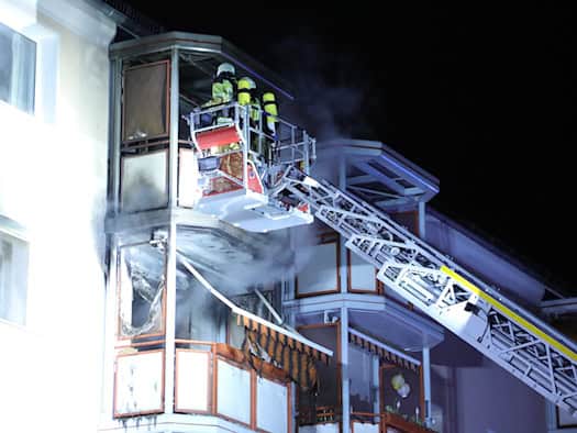 Feuerwehrleute in Schutzkleidung auf einer Drehleiter löschen einen Brand an einem Wohnungsbalkon im zweiten Obergeschoss eines Mehrfamilienhauses bei Nacht. Rauch steigt auf, Teile der Balkonverkleidung und Markise sind sichtbar verkohlt.