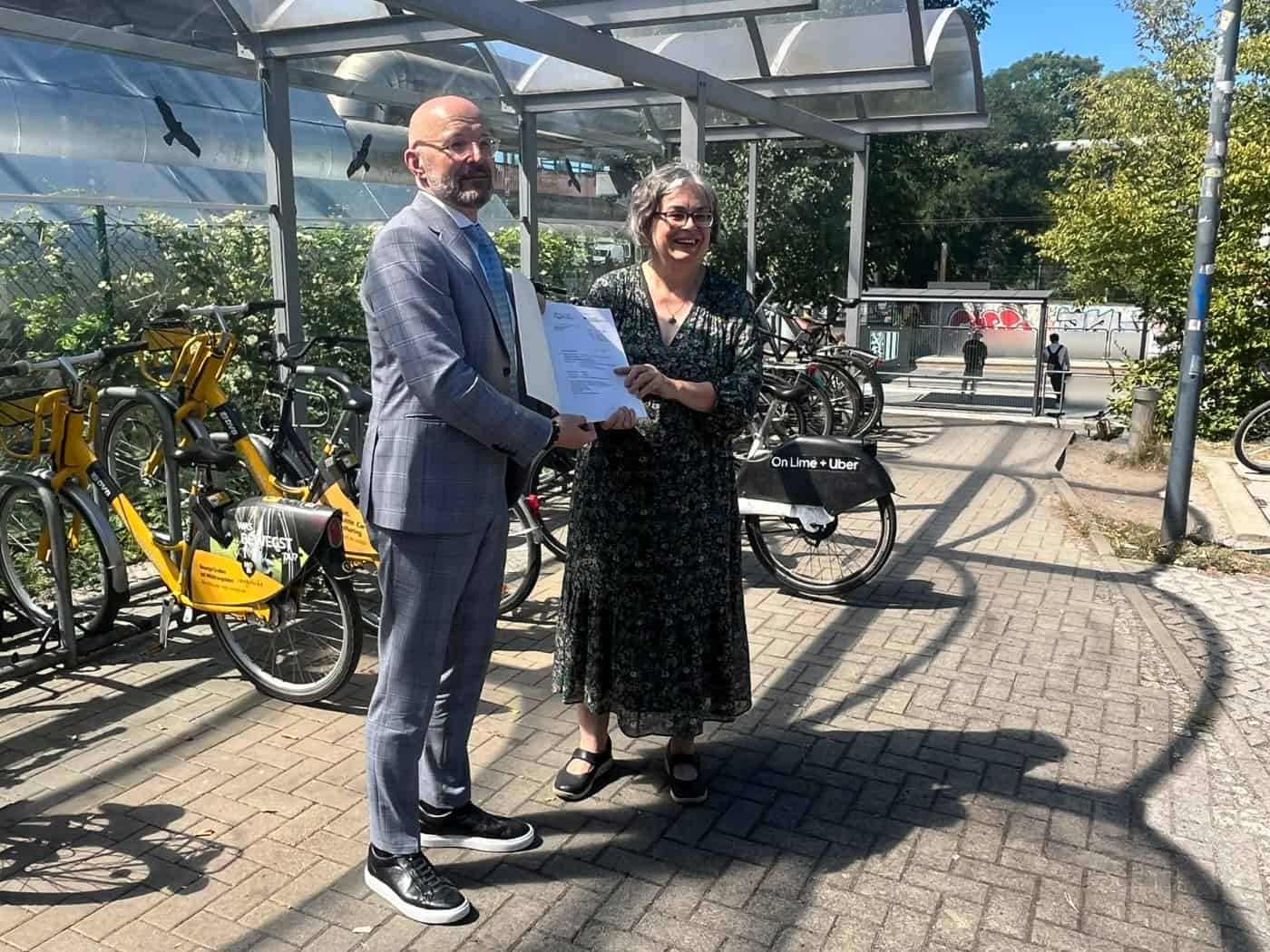 Thomas Kralinski vom Bundesumweltministerium im Anzug übergibt Dresdens Umweltbürgermeisterin Eva Jähnigen im Musterkleid einen Förderbescheid unter einem Fahrradunterstand. Im Hintergrund stehen gelbe MOBIbikes.