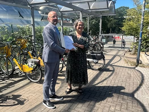 Thomas Kralinski vom Bundesumweltministerium im Anzug übergibt Dresdens Umweltbürgermeisterin Eva Jähnigen im Musterkleid einen Förderbescheid unter einem Fahrradunterstand. Im Hintergrund stehen gelbe MOBIbikes.
