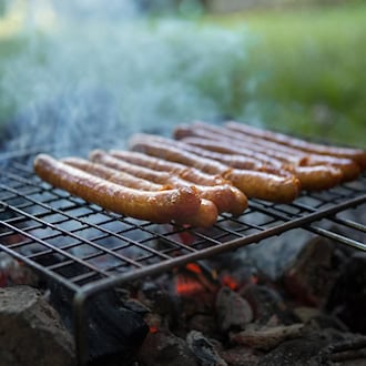 Nahaufnahme von mehreren braun gebratenen Würstchen auf einem Grillrost über glühenden Kohlen. Leichter Rauch steigt auf, im unscharfen Hintergrund liegt eine grüne Wiese im Freien.