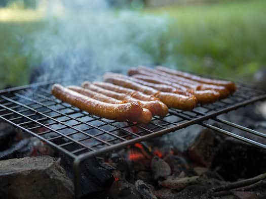 Nahaufnahme von mehreren braun gebratenen Würstchen auf einem Grillrost über glühenden Kohlen. Leichter Rauch steigt auf, im unscharfen Hintergrund liegt eine grüne Wiese im Freien.