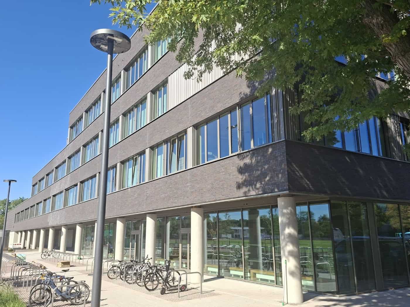 Das moderne Gebäude des Gymnasiums LEO in Dresden mit dunkler Klinkerfassade, großen Glasfenstern, Säulen im Erdgeschoss und Fahrradständern im Vordergrund bei sonnigem Wetter.
