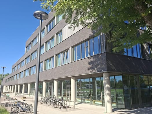 Das moderne Gebäude des Gymnasiums LEO in Dresden mit dunkler Klinkerfassade, großen Glasfenstern, Säulen im Erdgeschoss und Fahrradständern im Vordergrund bei sonnigem Wetter.