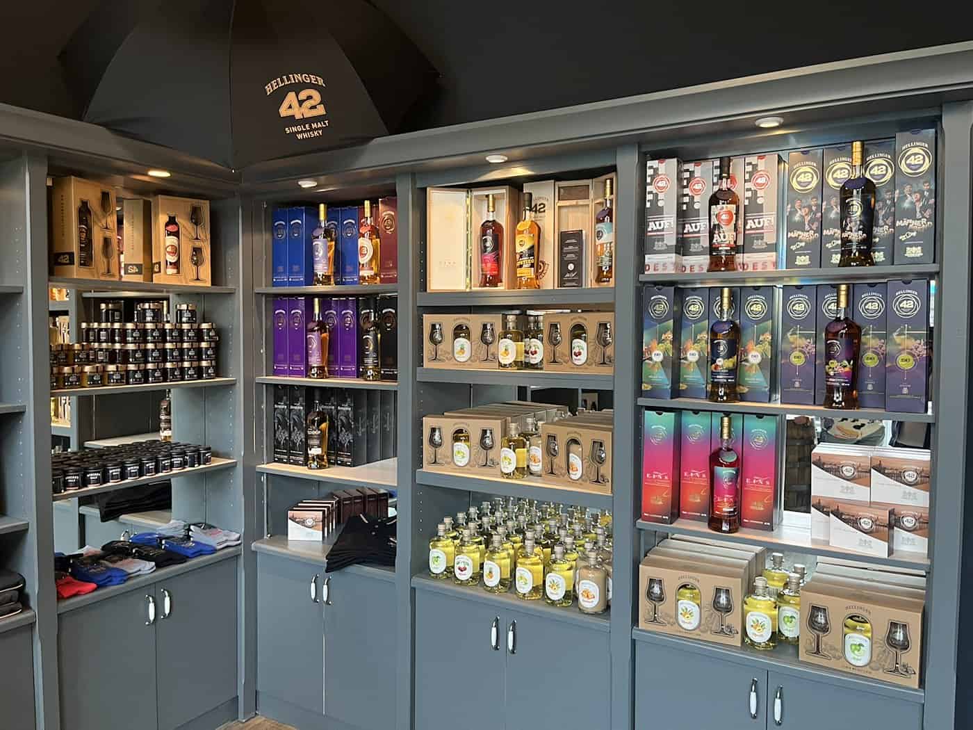 Gut gefüllte graue Holzregale im neuen Verkaufsraum der Dresdner Whisky Manufaktur. Zu sehen sind Flaschen des Whiskys Hellinger 42, Geschenksets, Gläser sowie ein schwarzer Schirm mit dem Markenlogo oben links.