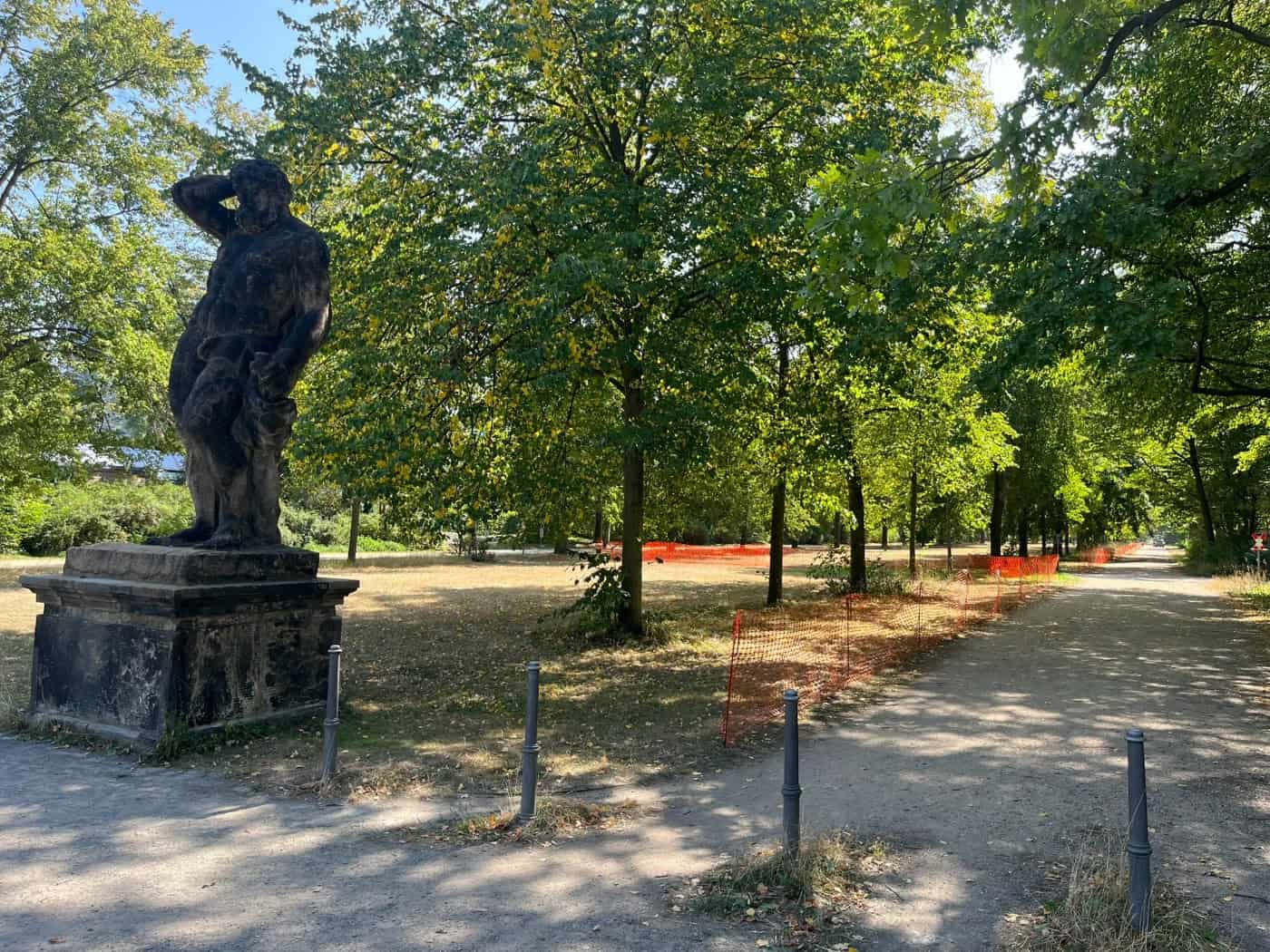 Eine historische Sandsteinstatue auf einem Sockel im Vordergrund am Eingang zur Herkulesallee im Großen Garten Dresden. Der von Linden gesäumte Parkweg ist dahinter mit orangenen Schutzzäunen abgesperrt.