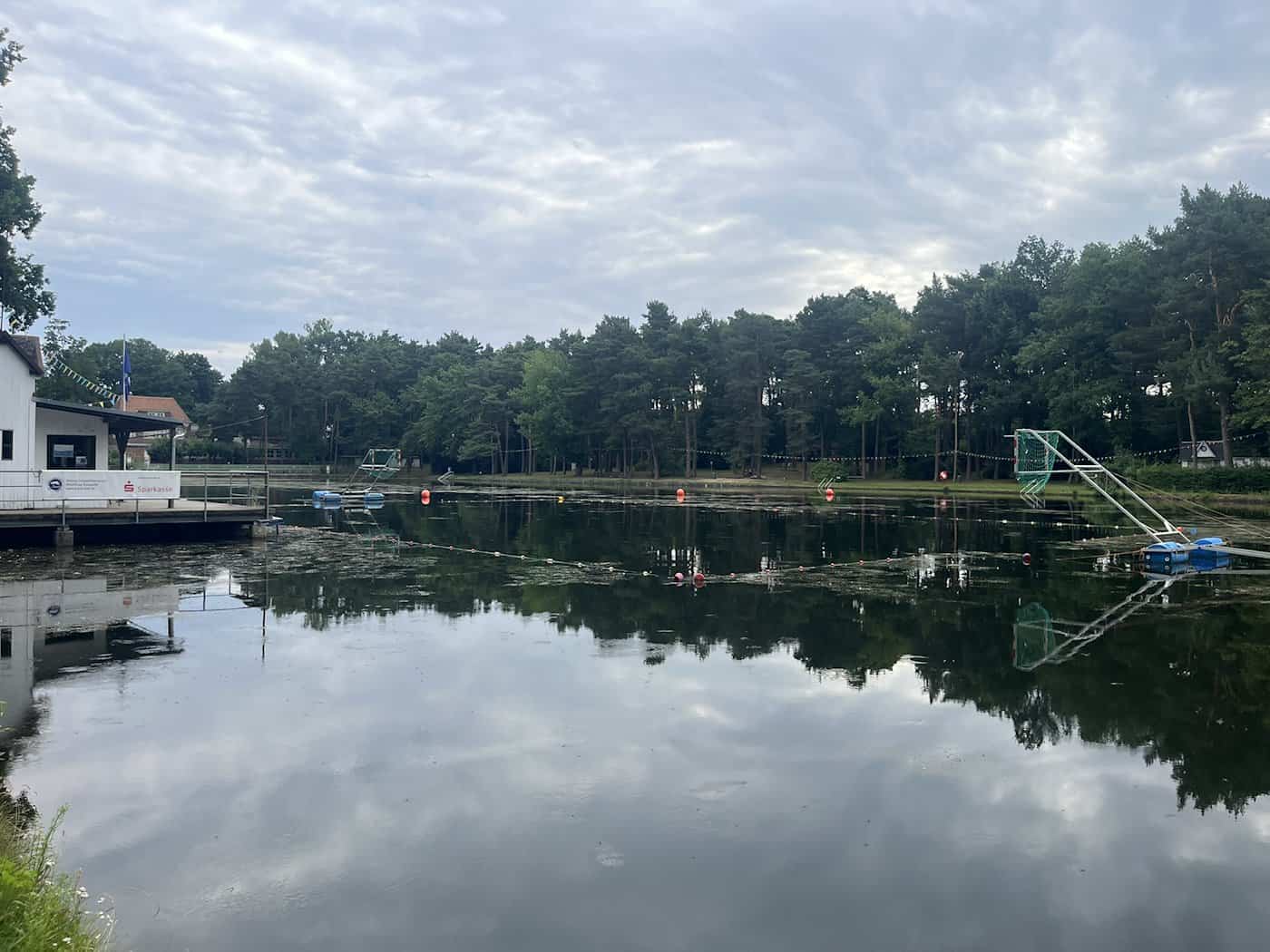 Ruhige Wasserfläche der Badestelle Weixdorf mit einer rot-weißen Begrenzungsleine im grünlichen Wasser, umgeben von dichten Bäumen und hellem Wolkenhimmel.