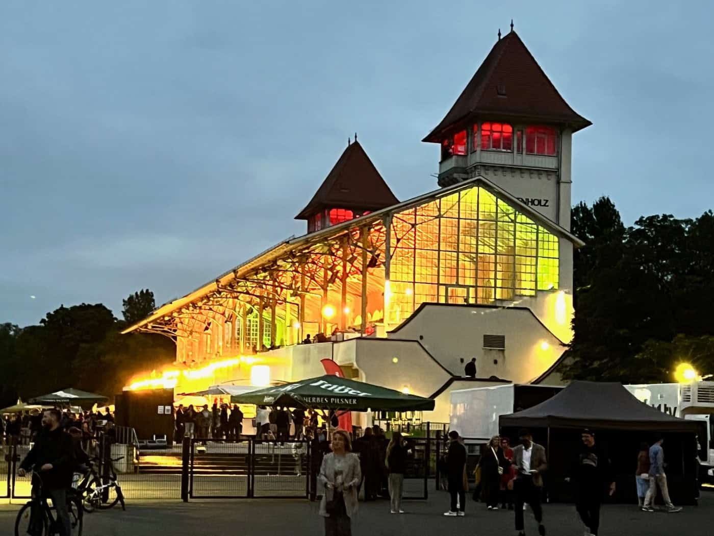 Abendliche Stimmung am Partyrenntag: Die beleuchtete Tribüne der Galopprennbahn Scheibenholz mit ihren markanten Türmen und lebhaftem Treiben vor dem Gebäude.