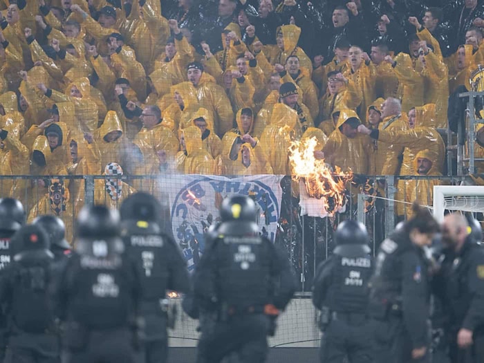 Dynamo Dresden-Fans im K-Block während der Ausschreitungen gegen Hertha BSC. Im Vordergrund sichern zahlreiche Polizeikräfte den Innenraum, während im Fanblock ein brennender Gegenstand zu sehen ist.