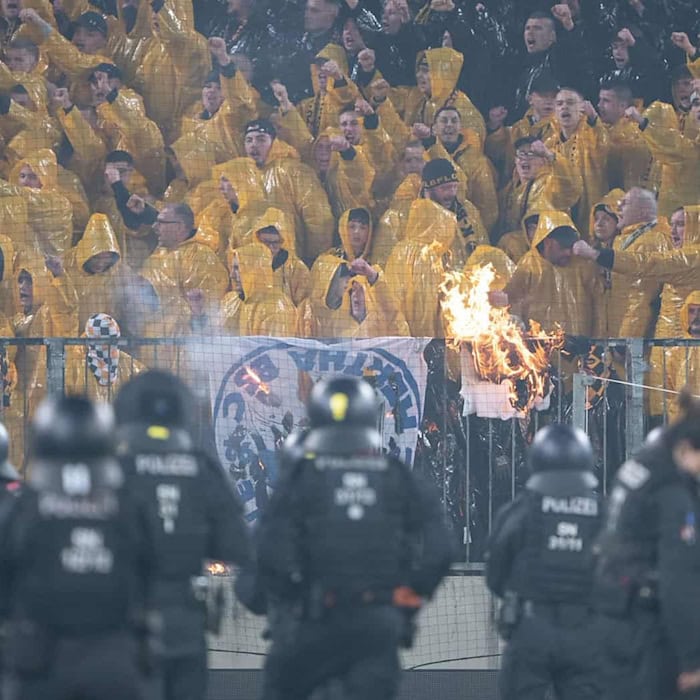 Dynamo Dresden-Fans im K-Block während der Ausschreitungen gegen Hertha BSC. Im Vordergrund sichern zahlreiche Polizeikräfte den Innenraum, während im Fanblock ein brennender Gegenstand zu sehen ist.