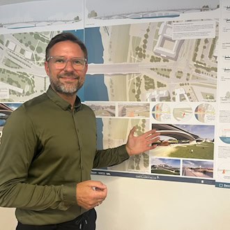 Ein Mann mit Brille und grünem Hemd steht vor einer Plakatwand mit Entwürfen zur neuen Carolabrücke in Dresden. Mit geöffneter Hand deutet er auf eine Architektur-Visualisierung der Brücke.