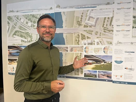 Ein Mann mit Brille und grünem Hemd steht vor einer Plakatwand mit Entwürfen zur neuen Carolabrücke in Dresden. Mit geöffneter Hand deutet er auf eine Architektur-Visualisierung der Brücke.