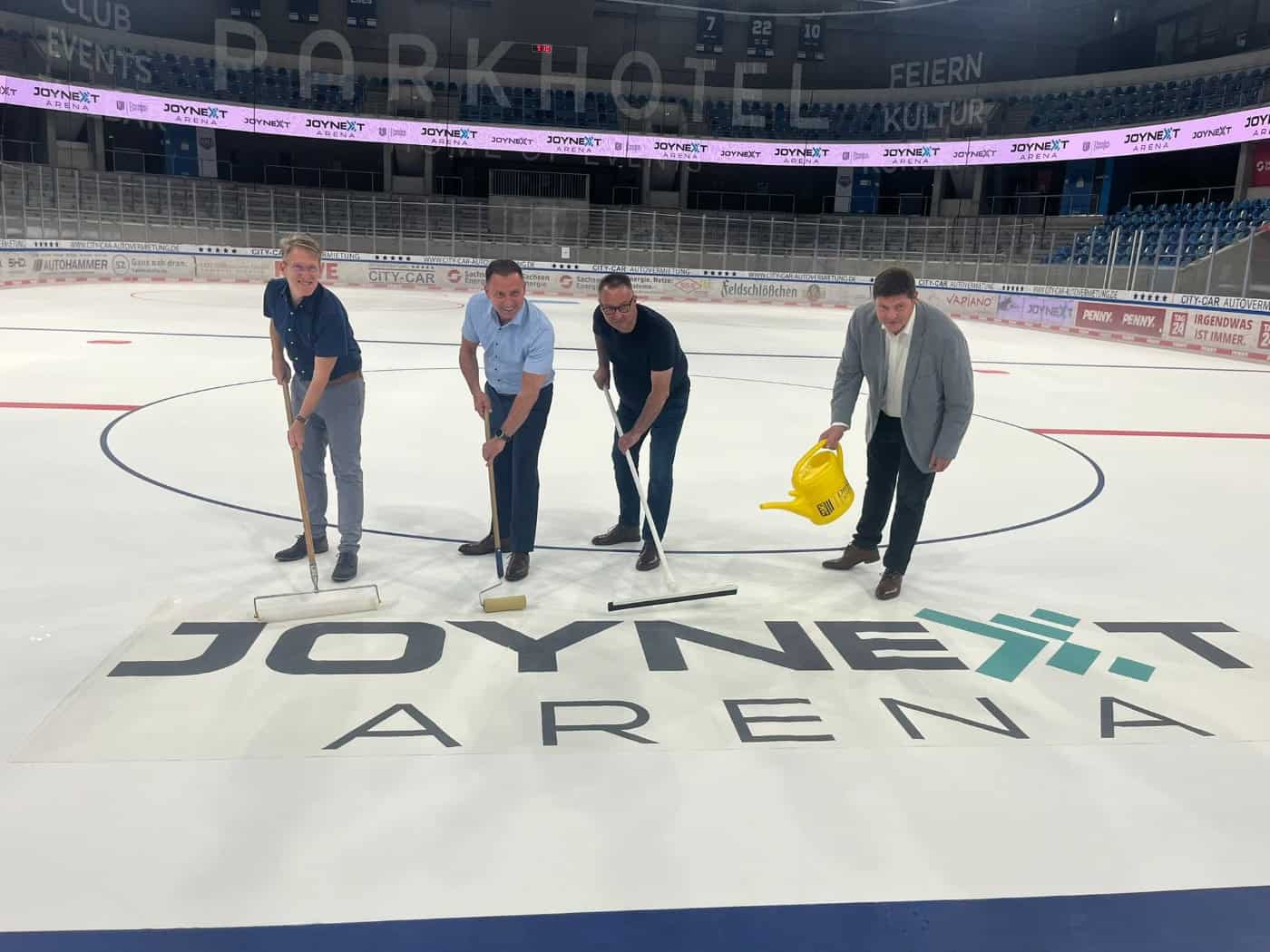 Vier Männer auf der Eisfläche einer Sporthalle bringen gemeinsam ein großes „Joynext Arena“-Logo auf dem Eis an. Sie nutzen dafür Walzen, Abzieher und eine gelbe Gießkanne. Im Hintergrund ist die leere Zuschauertribüne der Eishalle zu sehen.