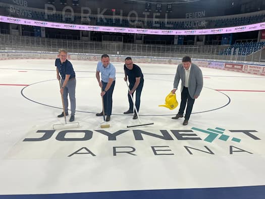 Vier Männer auf der Eisfläche einer Sporthalle bringen gemeinsam ein großes „Joynext Arena“-Logo auf dem Eis an. Sie nutzen dafür Walzen, Abzieher und eine gelbe Gießkanne. Im Hintergrund ist die leere Zuschauertribüne der Eishalle zu sehen.
