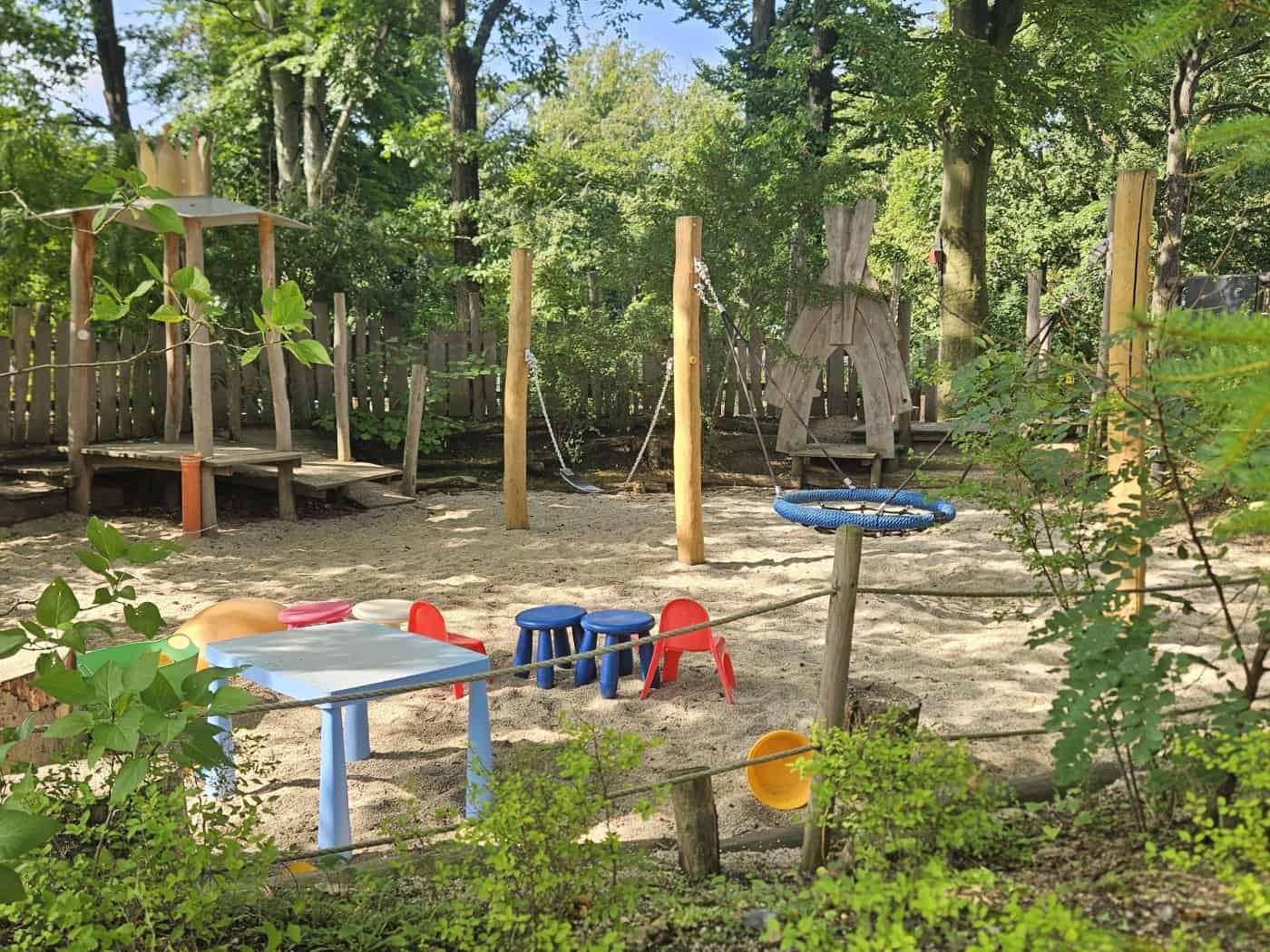 Ein naturnaher Spielplatz im Schatten hoher Bäume mit Sandfläche, bunten kleinen Plastikstühlen und einem blauen Tisch. Im Hintergrund stehen Holzspielgeräte wie eine Schaukel, ein Holzturm und Tipis, umgeben von dichtem Grün.
