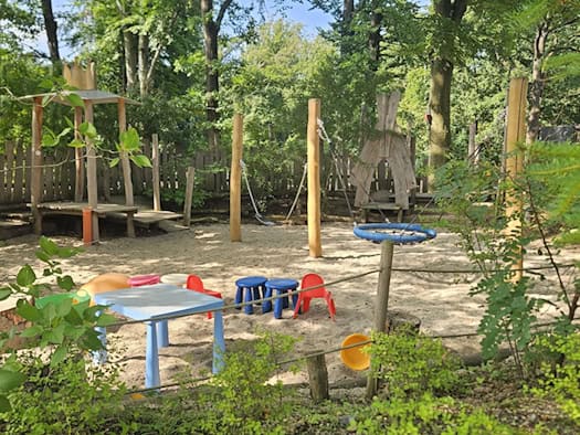 Ein naturnaher Spielplatz im Schatten hoher Bäume mit Sandfläche, bunten kleinen Plastikstühlen und einem blauen Tisch. Im Hintergrund stehen Holzspielgeräte wie eine Schaukel, ein Holzturm und Tipis, umgeben von dichtem Grün.