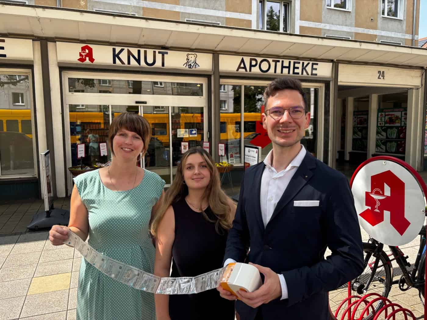 Romy Schneider, Jasmin Schmähl und Michael Gäbe stehen vor dem Eingang der Knut-Apotheke in Dresden und präsentieren ein langes Band maschinell verblisterter Medikamentenbeutel.